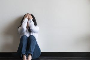 A woman sits against the wall with her hands covering her face. This could represent the pain that is addressed in online EMDR in Florida. An online EMDR therapist can support you with online EMDR therapy in Florida. Contact us today!