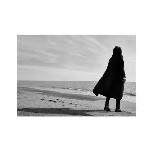 Person in dark coat standing alone on a quiet beach, looking out at the ocean under a cloudy sky
