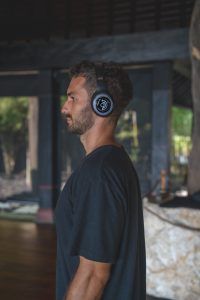 "Side profile of a man wearing black headphones with a circular logo, standing in a serene, open-air space with natural light filtering in. He appears calm and focused, possibly engaged in meditation or breathwork. The background features wooden elements and greenery, enhancing the tranquil atmosphere."