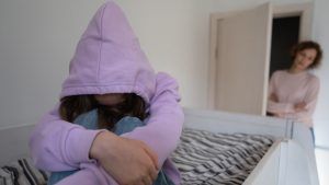 A teen sits with their hood over their face as their mother watches them. Online therapy for teens in Atlanta, GA can offer support for you and your teen from the comfort of home. Learn more about online therapy for teens by contacting an online therapist for teens in Atlanta, GA.